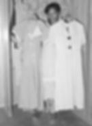 The image shows a woman proudly holding up two dresses she has made from feed sack cloth. In appearance, the dresses are plain button-down shirt dresses, knee-length and with short sleeves.