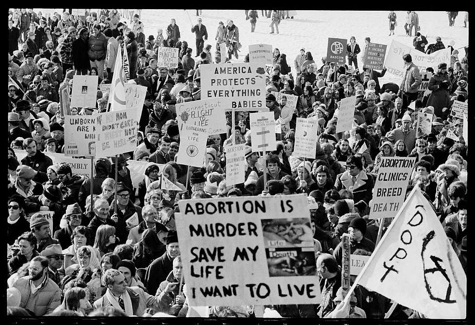 A black and white photograph of a large group of protestors carrying placards marked with anti-abortion slogans such as 'America Protects Everything but Babies'.
