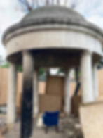 Weathered circular stone pavilion with a domed roof supported by columns, one blackened by fire damage. The interior looks abandoned, cluttered with debris and wooden boards blocking openings. A single blue chair sits inside on the dirt ground.