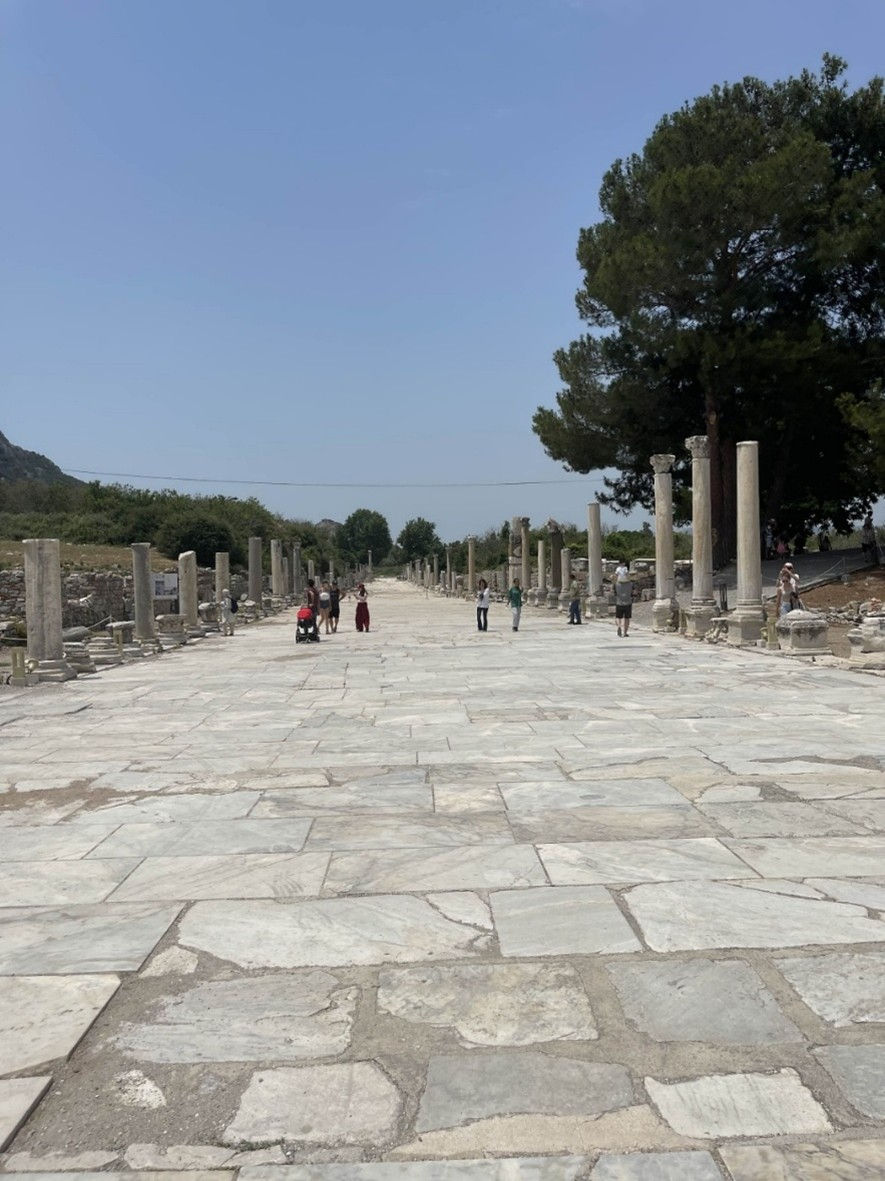 Photo shows the paved streets of Ephesus, with the paving stones various shades of sandy grave and chipped after centuries of use. The bright blue sky overlooks the seen as tourists walk up and down the street, with the remains of ancient columns on either side.