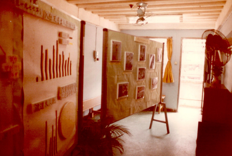 A small room lit by natural light from an open door on its far side. A large board with black and white photographs stands in the centre of the room.