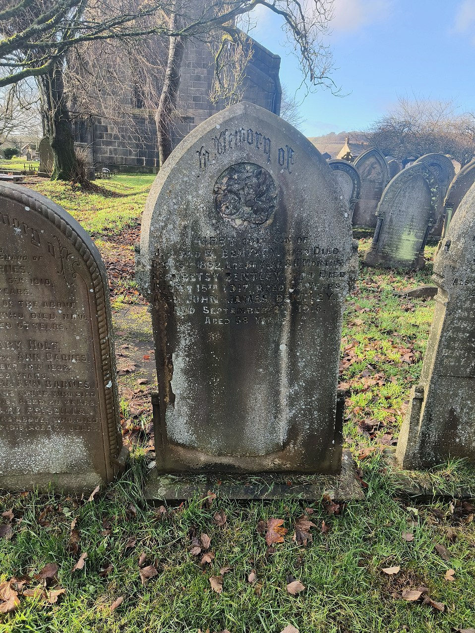 A photograph of a gravestone partially obscured by grey lichen. It is surrounded by rows of similar gravestones and there is a stone church in the background. The text is only partially legible but reads ‘in memory of’ at the top, with the name John James Bentley visible towards the middle of the headstone.