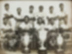 A group of men pose for a picture with three trophies on the ground. Five sit in the foreground and six other players stand behind them. All players are wearing boots, striped socks and a team shirt.