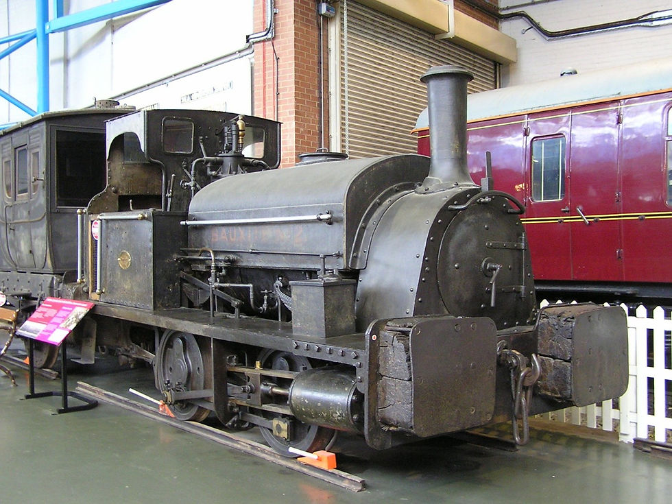 A small steam locomotive with four small wheels and large wooden buffers sits in the centre of the photograph. Its black paintwork is dull and filthy, as is the carriage behind it. In the background is a shiny maroon railway carriage, and a pair of industrial roller-doors.
