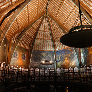 Photograph courtesy of The Oxford Union (CC BY 2.0.). A large room containing bookcases that are dwarfed by a grand ornate ceiling. The ceiling is made up of red coloured beams with yellow panels. Ornate paintwork decorates the walls while two brazier style chandeliers hang from the ceiling.