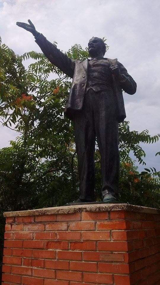 A tall bronze statue of a man with an outstretched arm, situated atop a red brick plinth.