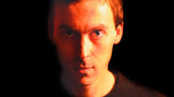Nick Land’s portrait with light reflecting on the left of his face with the other shadowed, showing a darker shade of red than on the lighter left-hand side. The setting is dark with no other objects visible.