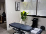 Chiropractic room with a blue adjustment table, potted plant, and cat poster. Window with blinds partially open, gray carpeted floor.