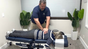 Dr. Frank in a blue shirt performs an adjustment on a woman lying face down on a table. Plants and blinds are visible in the room.
