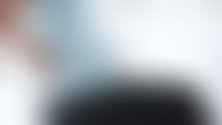 Pregnant woman in a blue shirt sits on a bed, holding her back in discomfort. The background features a white brick wall and pillows.