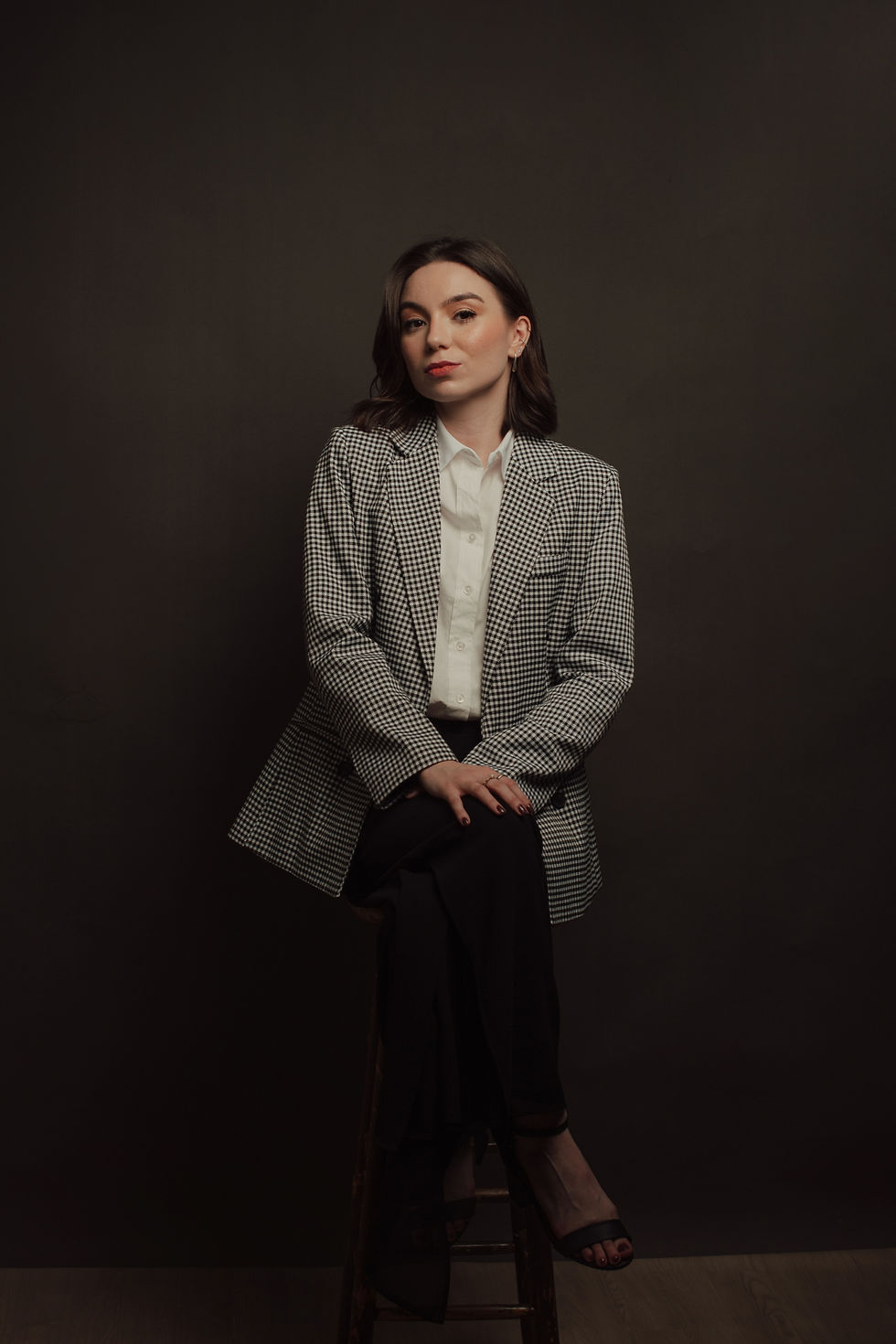 Retrato profissional de advogada sentada em um banco com blazer xadrez e fundo escuro.