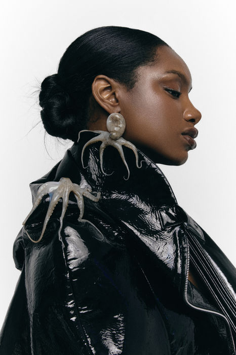 Model with radiant editorial makeup, sleek low bun hairstyle, black patent jacket, and sculptural octopus earrings for a high-fashion beauty photoshoot.