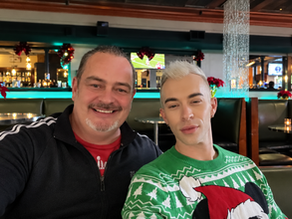 Two men posing for a selfie in a restaurant decorated for Christmas, one in a holiday sweater featuring Mickey Mouse in a Santa hat.