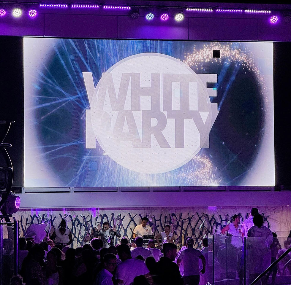 Crowd partying under purple lights with "White Party" sign, lively atmosphere, and people wearing white attire on a decorative backdrop.