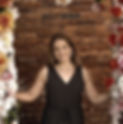 Woman in black dress smiles, standing between floral archways with colorful flowers. Brick wall background adds a rustic charm.