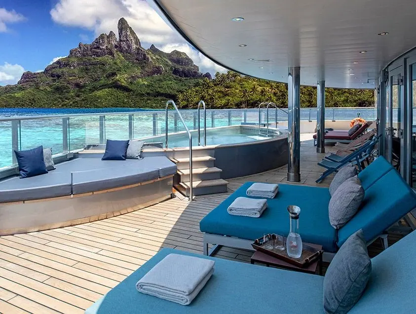 Cruise ship deck with loungers, a round pool, and blue cushions overlooking tropical island and turquoise water. Sunny, serene setting.