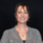 Portrait of a smiling woman wearing a patterned blouse, earrings, and a necklace, posed against a dark background.