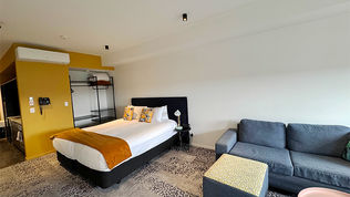 A hotel room with a king-size bed, a sofa, and an ottoman. The gold accent on the wall adds a stylish touch, and an open shelf holds towels and pillows. The room offers a spacious and comfortable setting for relaxation.