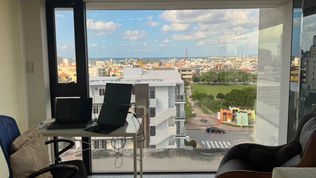 Office with a laptop on a desk in front of a large window. Cityscape and ocean visible outside on a clear day. Calm, organized space.