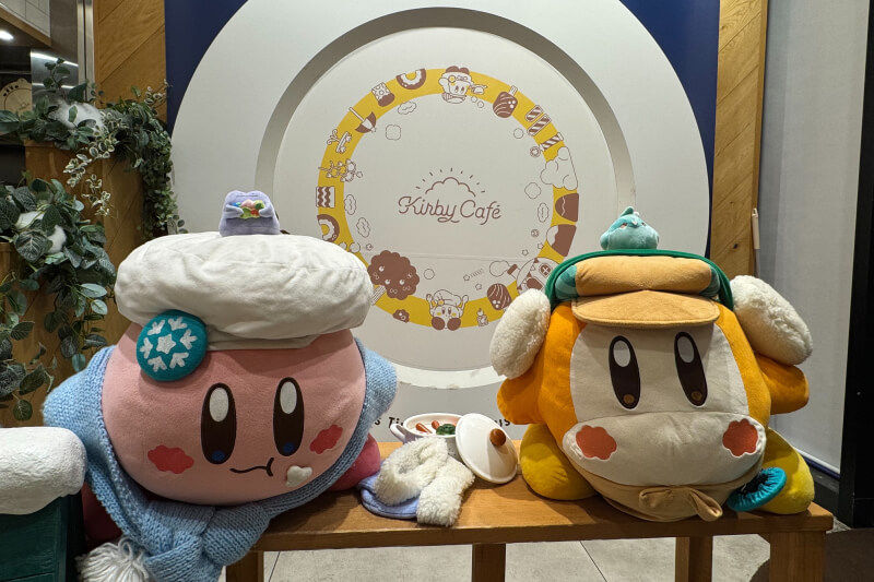 Plush Kirby and Waddle Dee in winter outfits sit on a bench at Kirby Café. A large decorative plate with the café logo is in the background.