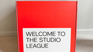 Red box on beige floor with text "Welcome to the Studio League" in bold black letters on a white label, against a plain wall background.