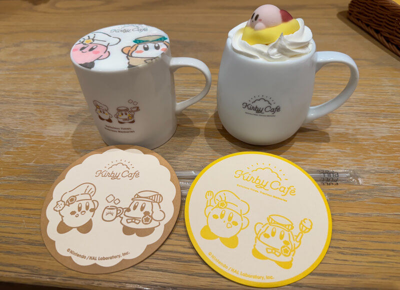 Two white mugs with Kirby designs, one topped with cream and a Kirby figure. Two coasters with Kirby Cafe logos and drawings on a table.