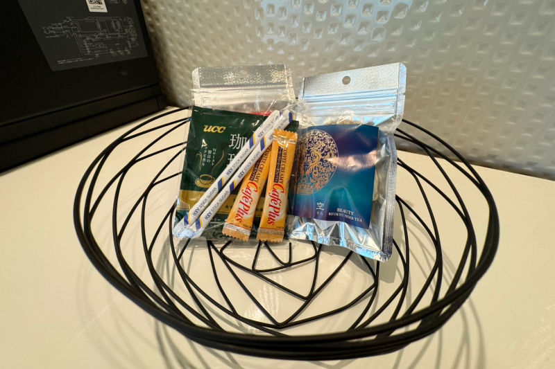 Basket with coffee, snacks, and tea packets on a white table. Background features a textured wall. Items include UCC coffee and Care Plus candies.