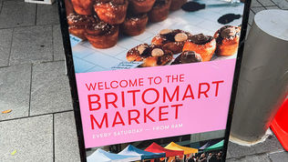 An A-frame sign placed on an asphalt sidewalk. The top half shows baked goods like muffins, while the bottom half displays a photo of colorful market tents. Bold text at the center reads, "WELCOME TO THE BRITOMART MARKET."