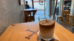 Iced coffee on a wooden table in a cozy cafe with a glass door view of street. A blurred figure stands near the entrance. Warm ambiance.