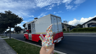 A soft serve ice cream with colorful toppings is held up toward the back of a parked ice cream truck on a residential street. The background features a clear blue sky and tree-lined sidewalks.