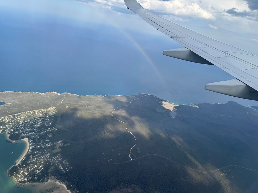 飛行機の窓から見たオーストラリアの海岸線。翼の下に青い海と陸地が広がり、海上には虹が見える。
