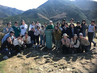 山の展望地で、20人ほどの若者が岩場に集まり、ピースサインで記念撮影している。背後に緑の山々が広がる。