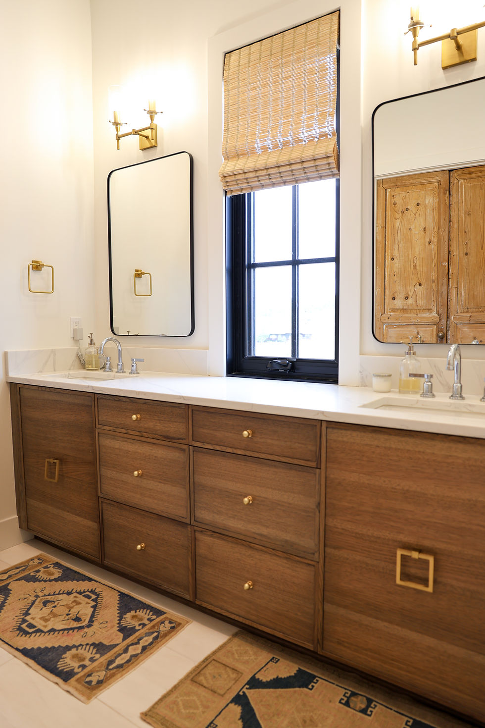 Bathroom with a custom wood vanity, two mirrors, and wicker blinds. Gold fixtures and neutral tones create a modern, serene atmosphere. Rugs on floor.