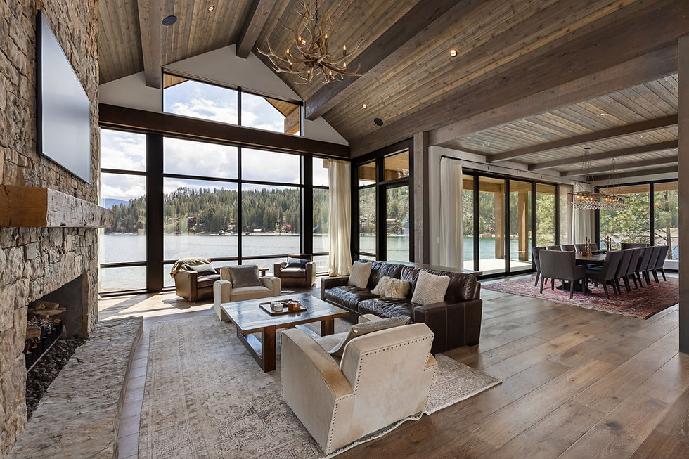 Luxury lakefront living room with stone fireplace, leather sofa, antler chandelier, and floor-to-ceiling windows overlooking water and pines