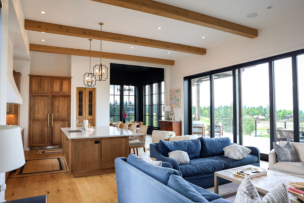 Bright open-plan living room and kitchen with blue sofas, wood beams, pendant lights, and large windows overlooking a green patio.