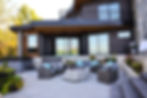 Modern patio with wicker seating, blue cushions, a stone fire pit, and glass doors. Surrounded by trees and set against a black house.