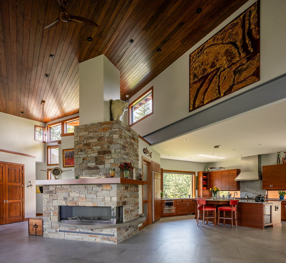Rustic open-plan kitchen and living room with stone fireplace, mounted goat, wood ceiling, red stools, and large windows.