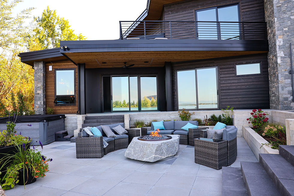 Modern stone-and-wood house patio with wicker seating around a lit fire pit, blue cushions, and a lake view through large windows.