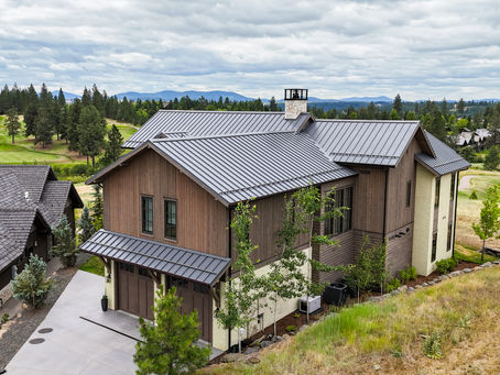 Modern custom North Idaho house with metal roof beside a golf course and pine trees under a cloudy sky, with mountains in the distance