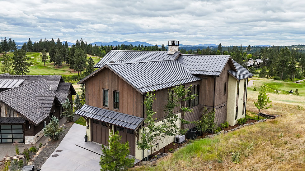Modern custom North Idaho house with metal roof beside a golf course and pine trees under a cloudy sky, with mountains in the distance
