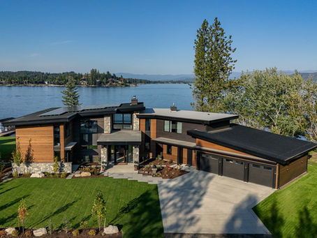 Modern lakeside house with black roof and wooden accents, surrounded by green lawn and trees, overlooking a calm blue lake and forested hills.