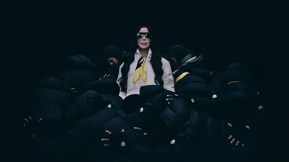Woman in sunglasses and white coat with yellow scarf stands amid hooded figures on a dark background, moody and dramatic.