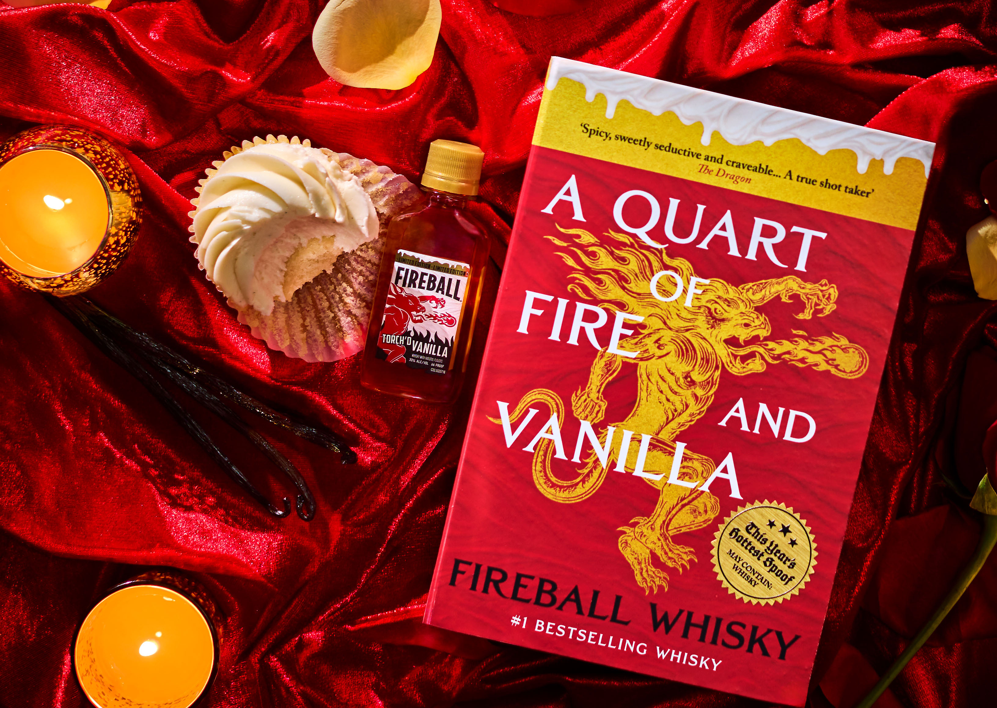Fireball Whisky book, mini bottle, and frosted cupcake on red satin with glowing candles, creating a bold, sultry display.
