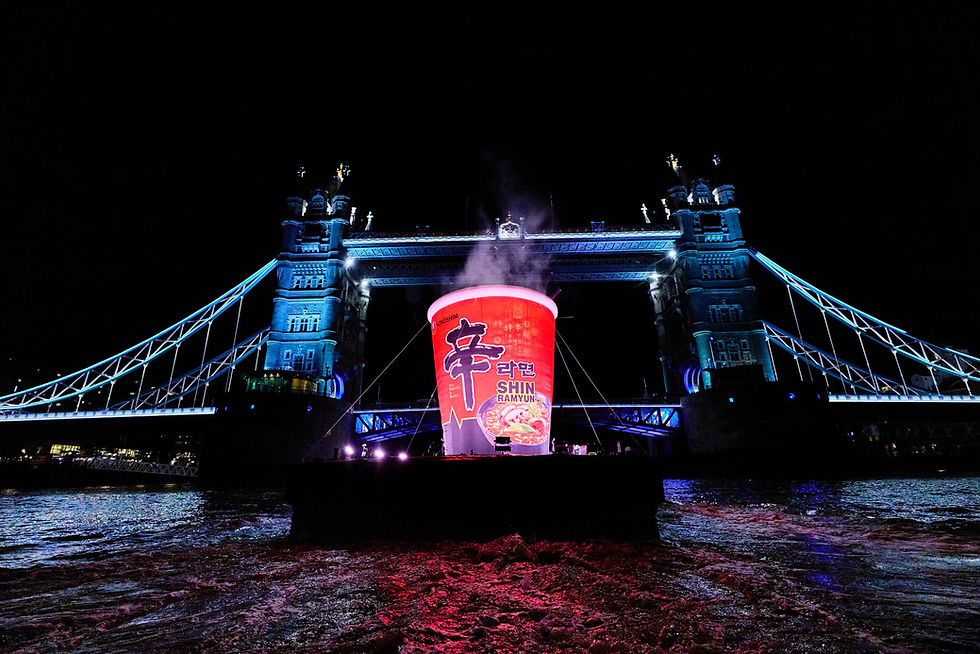 Tower Bridge at night lit blue, with a giant Shin Ramyun cup on a barge glowing pink over the dark river.