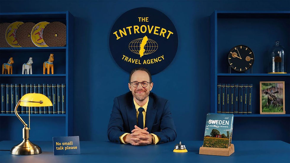 Smiling man in suit at blue desk in The Introvert Travel Agency, with yellow lamp, Sweden brochure and No small talk please sign.