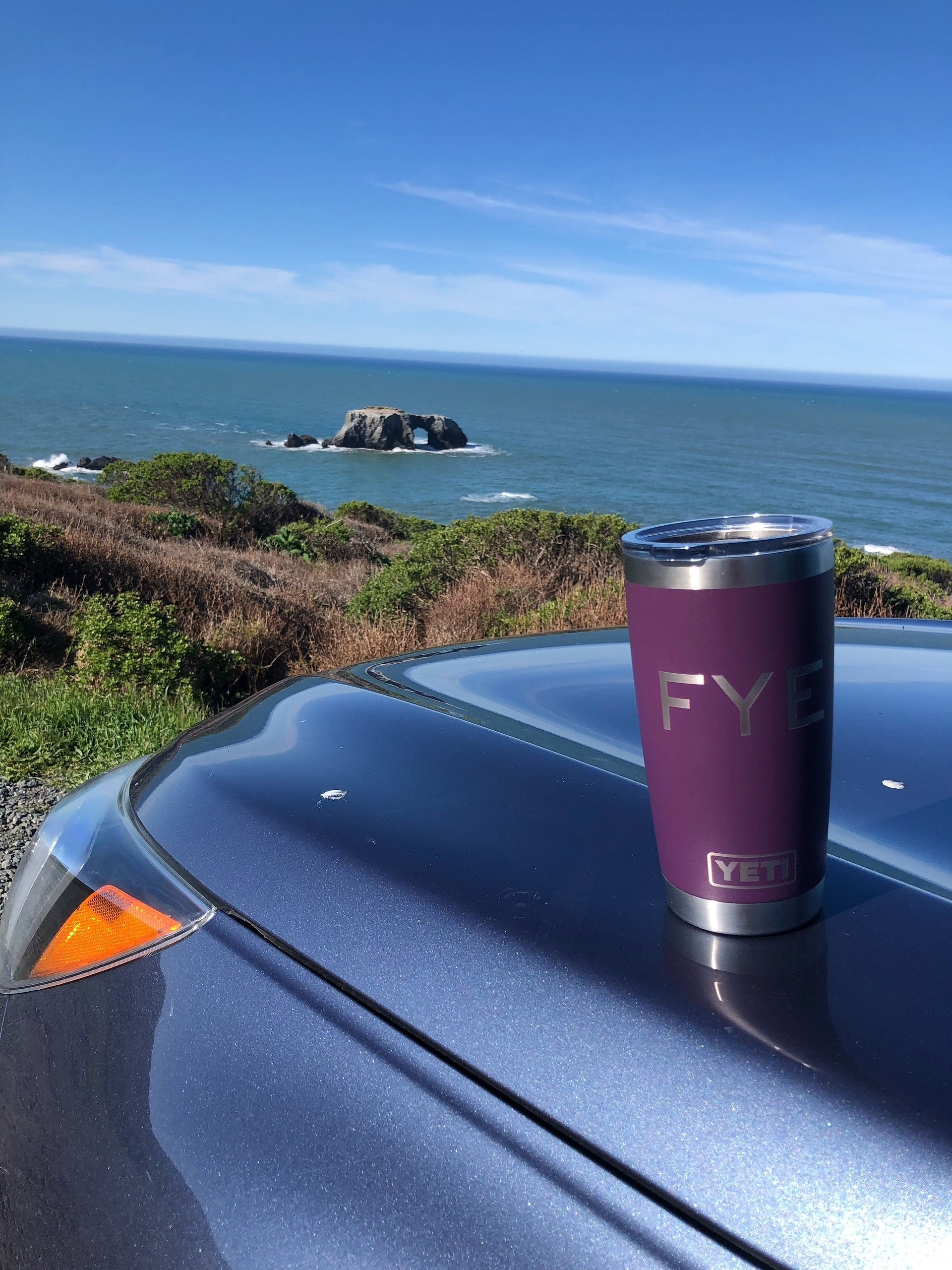 A blue Subaru Outback  parked by the ocean shore along CA hwy 1, with a purple Yeti mug on the hood. 