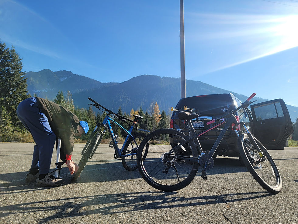 Paul Brightbill is bent over inflating the tire of a mountain bike. Another mountain bike is in the foreground, along with a car - while beautiful mountain rises behind them.