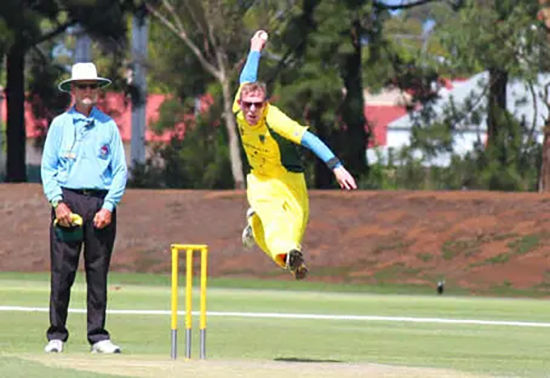 Australian bowler in action, about to bowl a ball. He is mid-flight about to release the ball, the umpire and stumps are also in view.