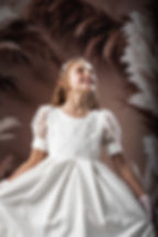 Young girl in white dress with lace, smiling, looking up. Brown feathered background creates elegant, joyful mood.
