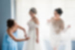 Bride in ornate white dress smiles, assisted by two women in blue and sequined dresses, against a window with blinds. Joyful mood.